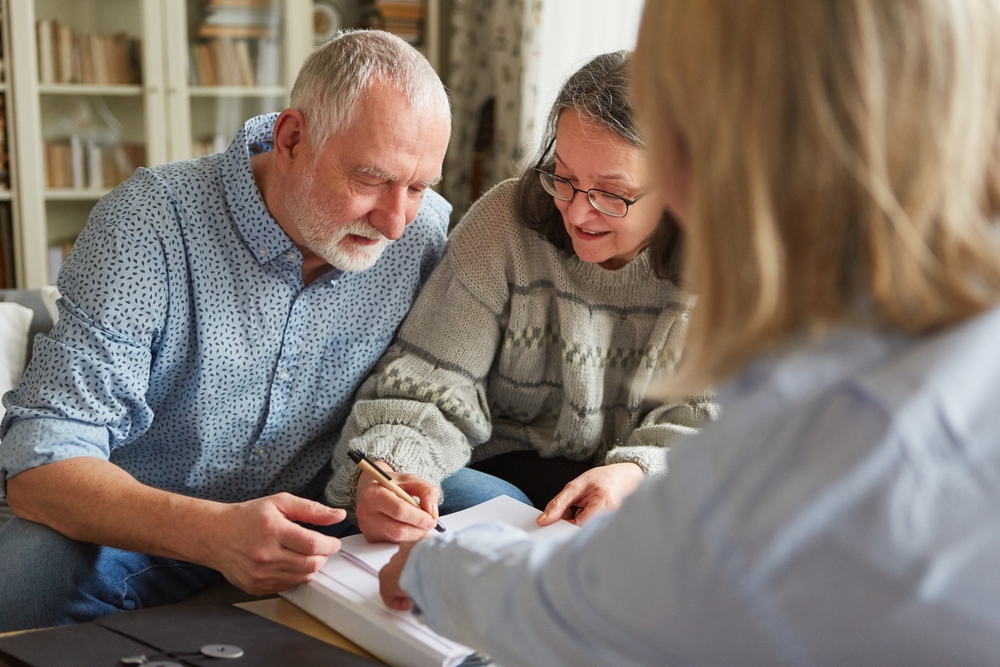 Older white couple signing paperwork with blurred out back of head pointing to beneficiary paperwork