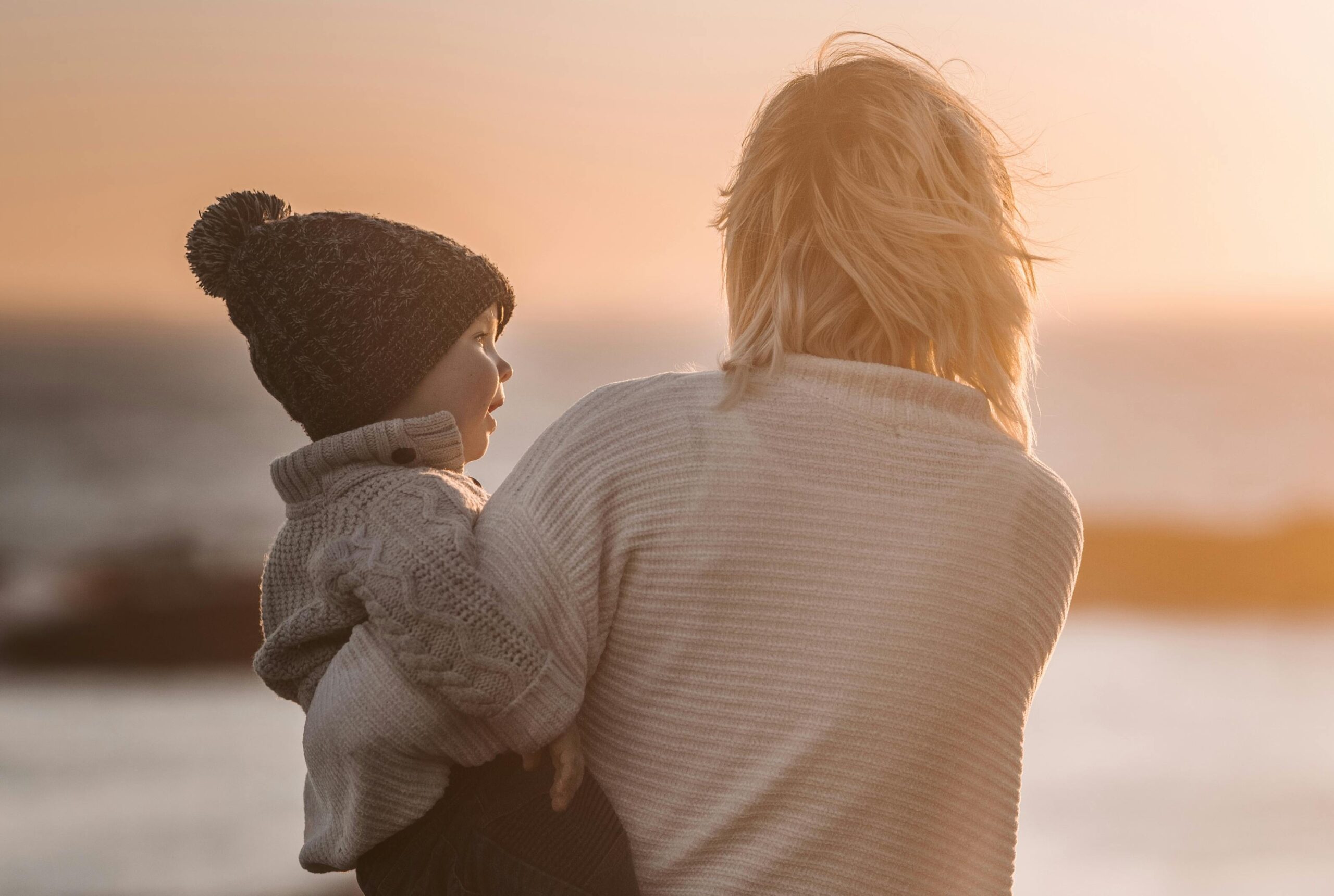 A mother holding her young child while looking out at a sunset. The child is wearing a knit sweater and a wool hat with a pom-pom, and the mother is wearing a light sweater. They both have their backs turned, gazing toward the horizon over a body of water.