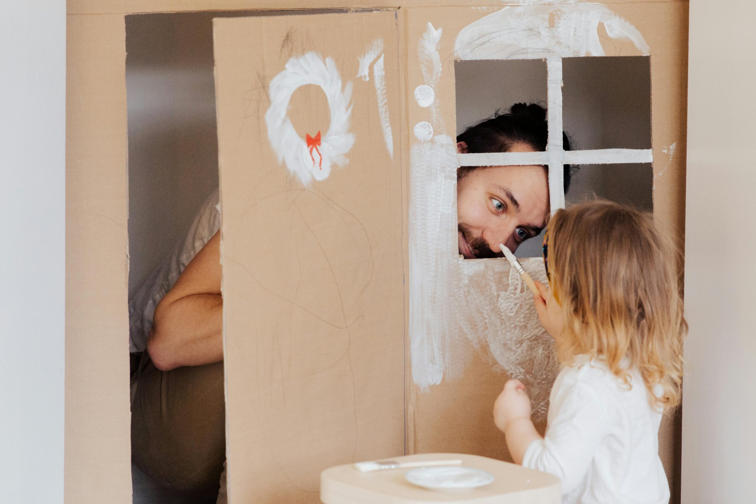 A young daughter painting on a cardboard house while her father playfully peeks through a cut-out window. The girl is focused on painting with a brush, and the cardboard house is decorated with white paint marks.