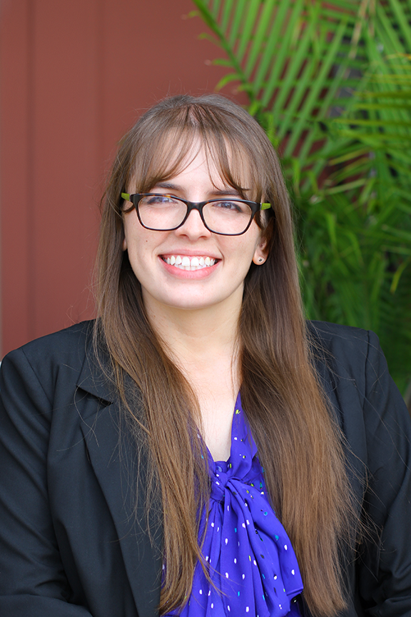 A portrait of Amy Parker, an attorney at Stimpson & Associates P.C., with long brown hair, wearing glasses, a black blazer, and a purple blouse. She is smiling and sitting in front of a background with green plants.