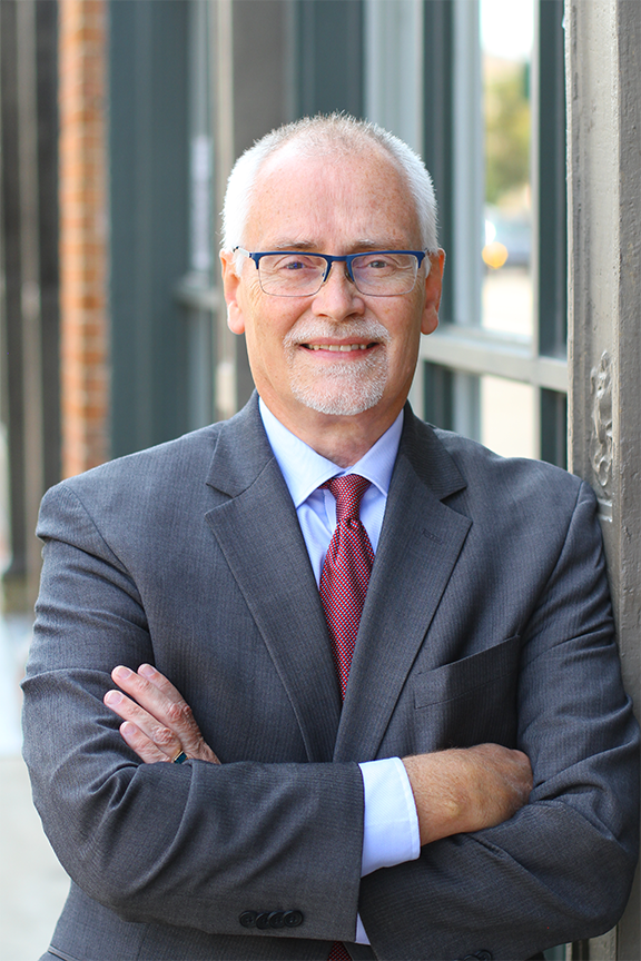 A portrait of David Lacasse, an attorney at Stimpson & Associates, wearing a gray suit, red tie, and glasses, smiling confidently with his arms crossed. He has short white hair and a light beard, standing in front of a building with large windows.