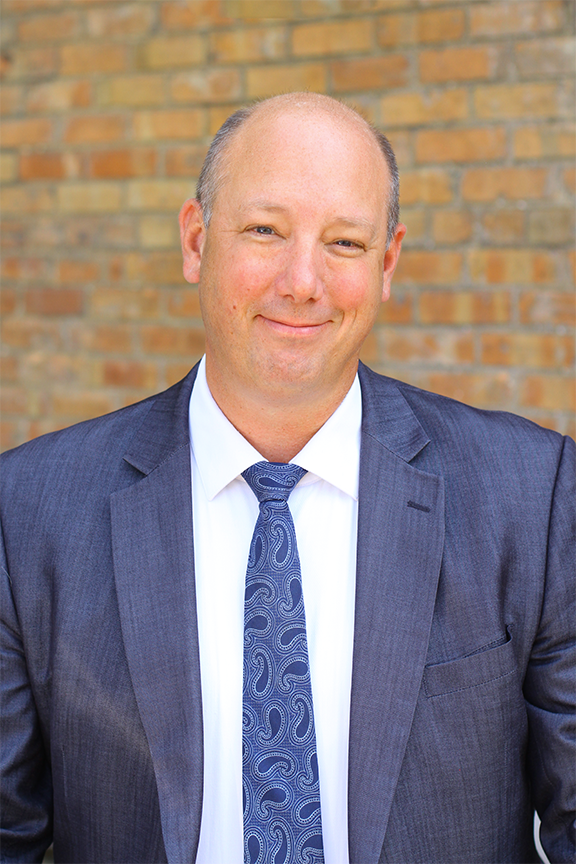 A portrait of David Stimpson, the Managing Partner of Stimpson & Associates, wearing a gray suit, white shirt, and blue patterned tie, smiling in front of a brick wall background.