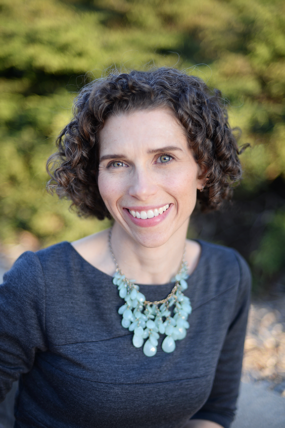 A portrait of Jessica Kelly, an attorney at Stimpson & Associates P.C., with short, curly hair, wearing a gray top and a statement necklace with light blue beads. She is smiling brightly and sitting outdoors with a blurred green background.